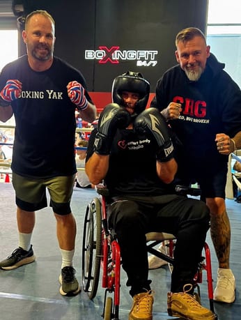 Two coaches standing either side of an adaptive boxer in a wheelchair inside a boxing ring