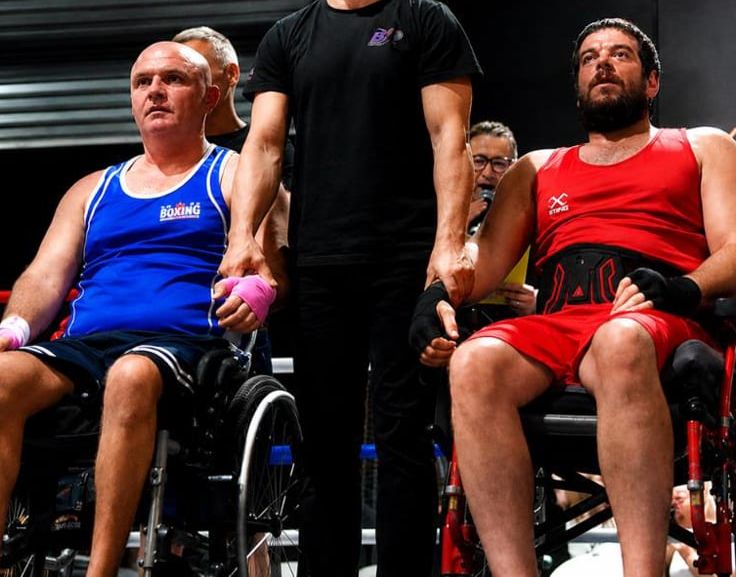 Two adaptive boxers in wheelchairs alongside their coach at a boxing event