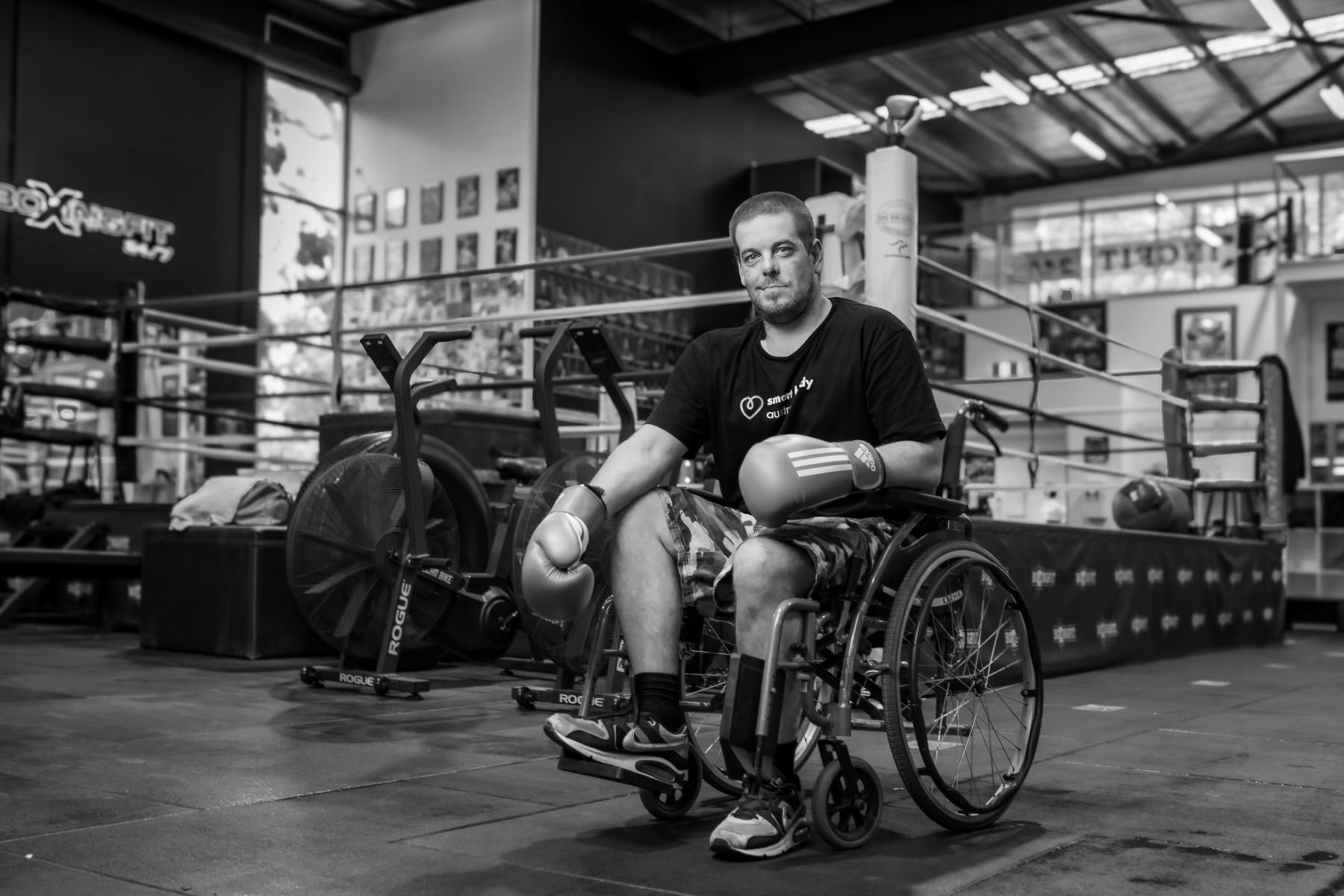 Black-and-white portrait of a boxer seated in his wheelchair beside the ring, gloves on his lap