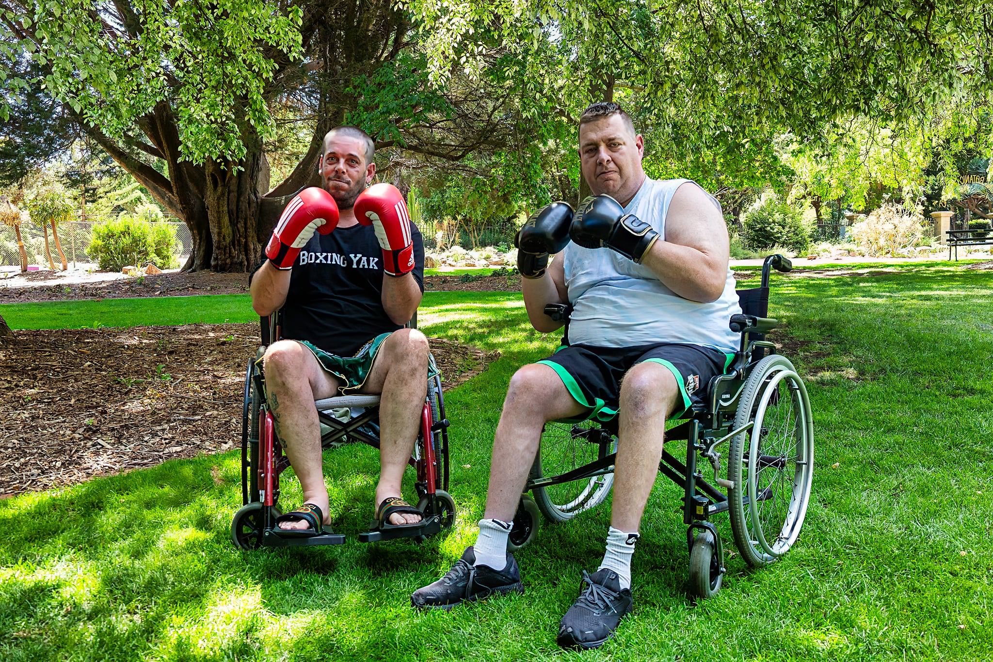 Two wheelchair boxers pose side by side under a large tree in a sunlit park